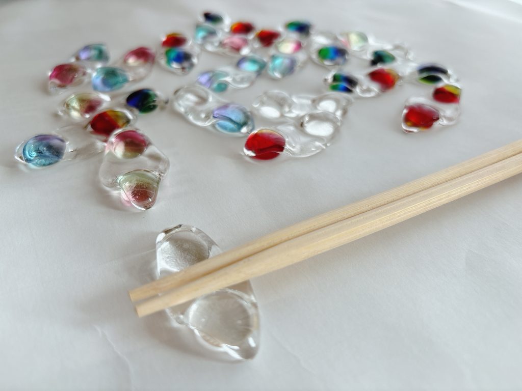 白い紙の上に並べられたカラフルなガラスの枝豆箸置き。手前にはお箸が載せられ、二粒の豆が並んだ新しい形状が見える。