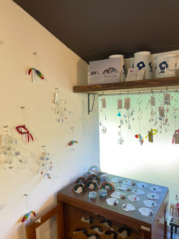 田川市の委託販売先「Comorebi」の店内。白い壁に虹色のガラス作品「虹魚」がディスプレイされ、棚には虹のモチーフのインテリア雑貨が並んでいる様子。