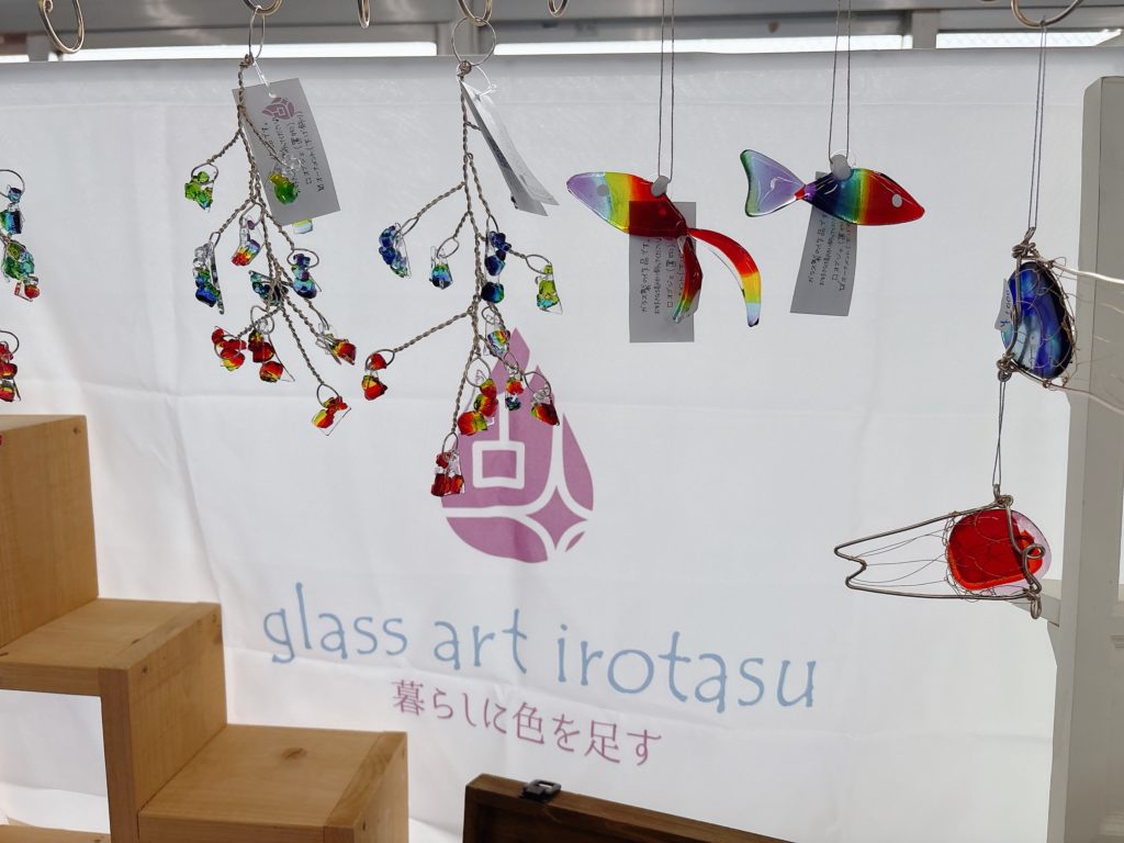 虹色のガラスオーナメントと金魚が吊り下げられたディスプレイ。背景に「glass art irotasu 暮らしに色を足す」のロゴが写る。