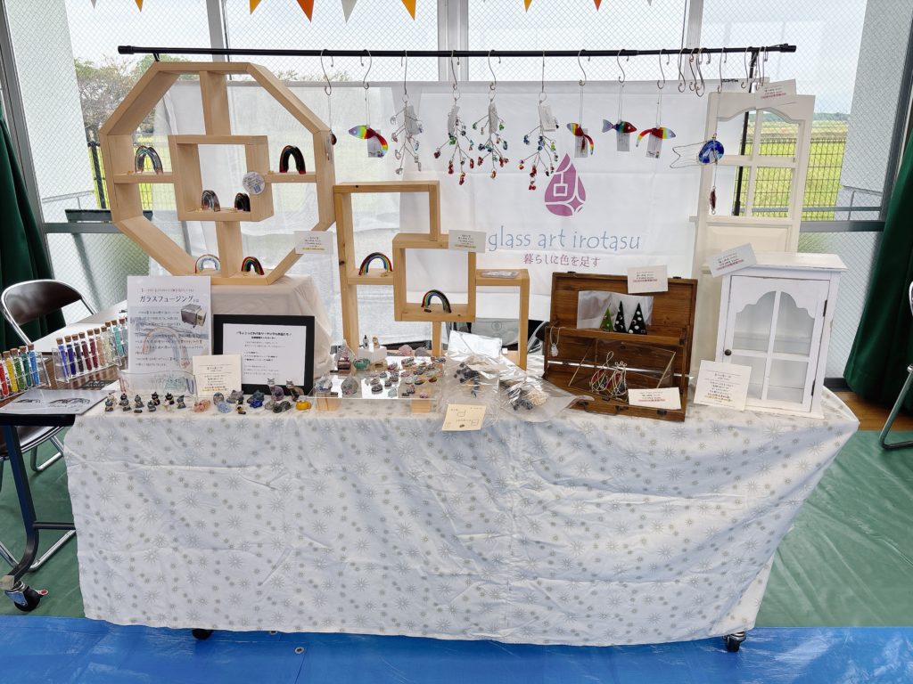 たちあらいマルシェでのイロタスのブース全景。虹のオブジェや吊り下げ型のガラス作品が並び、中央にロゴ入り布が掛かっている。