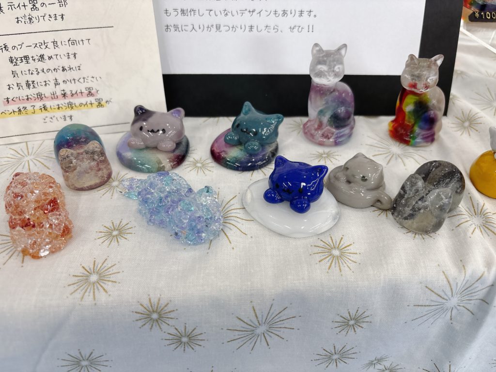 様々なポーズと色のガラス製猫オブジェがテーブルに並ぶ。虹色、透明、パステルなど多彩な仕上がり。