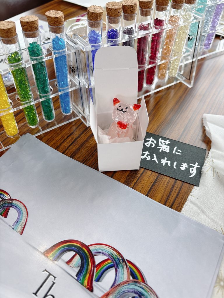 色とりどりのガラス粒が入った試験管と、透明なピンク猫の作品。黒い札に「お箱にお入れします」の文字。